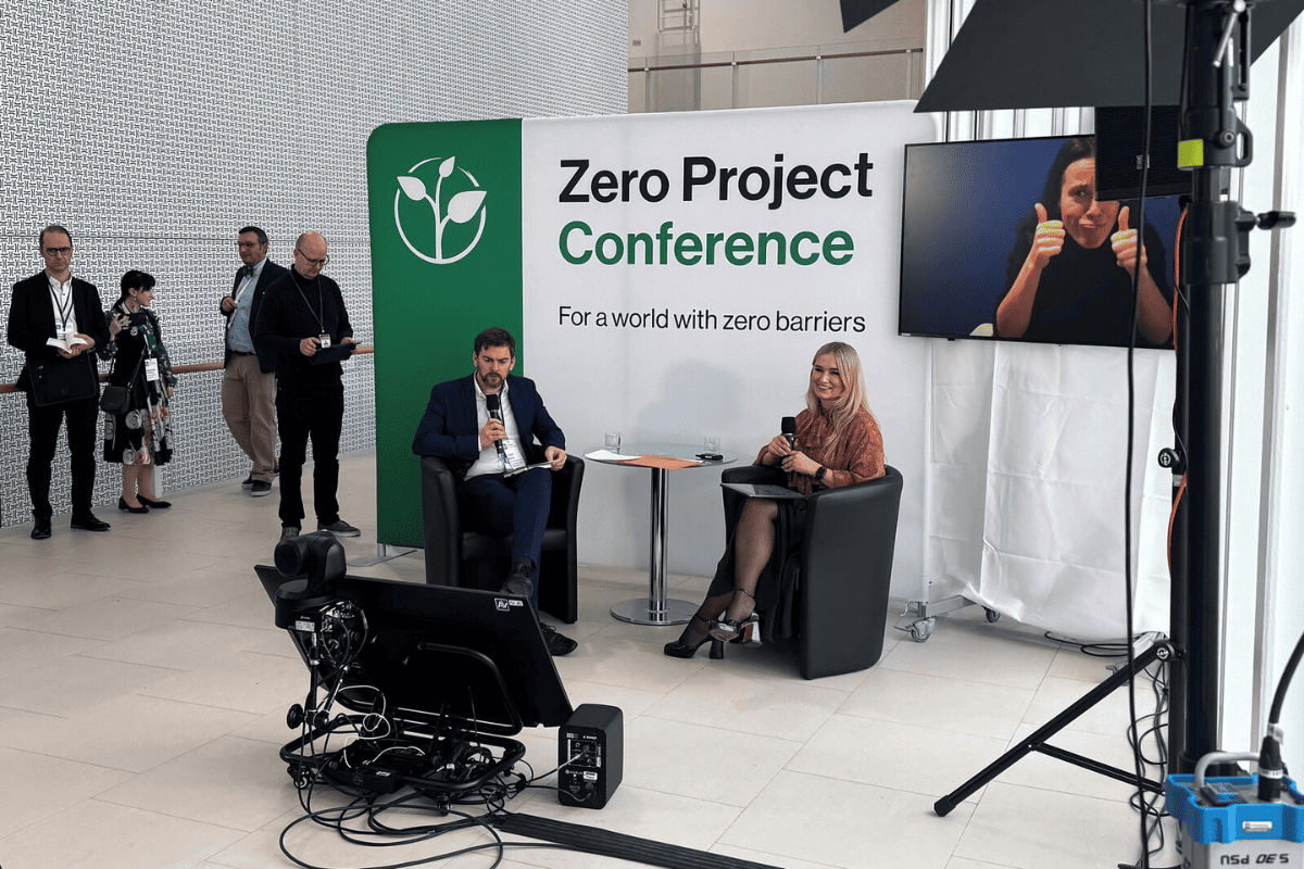 Dwie osoby siedzą w fotelach podczas rozmowy na scenie Zero Project Conference. W tle ścianka z napisem „Zero Project Conference – For a world with zero barriers”. Obok ekran z tłumaczeniem migowym.